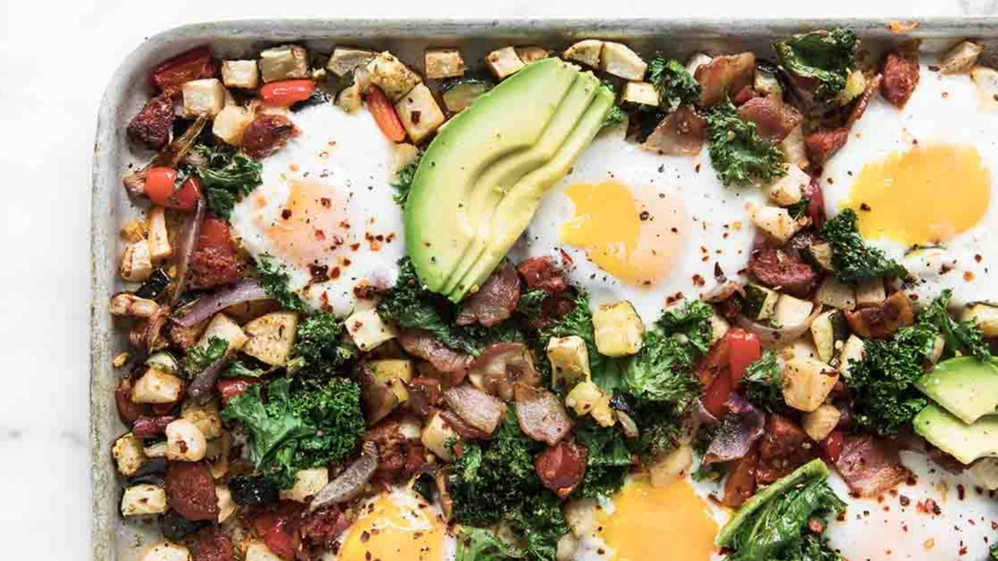 Overhead view of a baked breakfast hash on a sheet pan, featuring sunny-side-up eggs, chopped kale, avocado slices, red onion, and diced vegetables with a sprinkle of red pepper flakes.