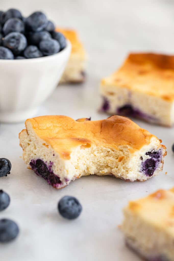 one of the blueberry cheesecake bars bitten through beside a bowl of blueberries
