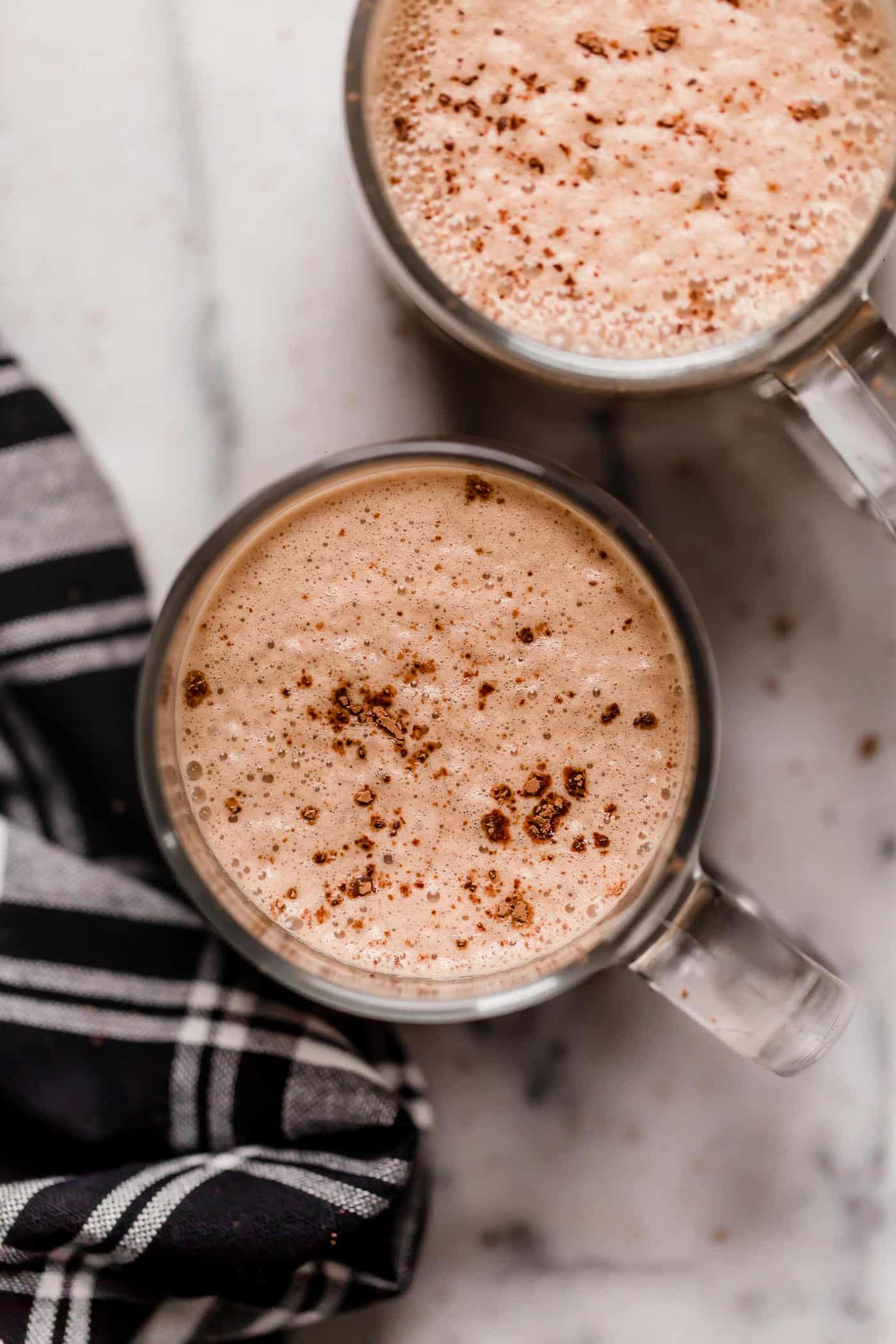 overhead shot of 2 clear glass mugs of bulletproof peppermint mocha with cocoa powder sprinkled on top atop a white marble countertop next to a black and white checkered linen napkin