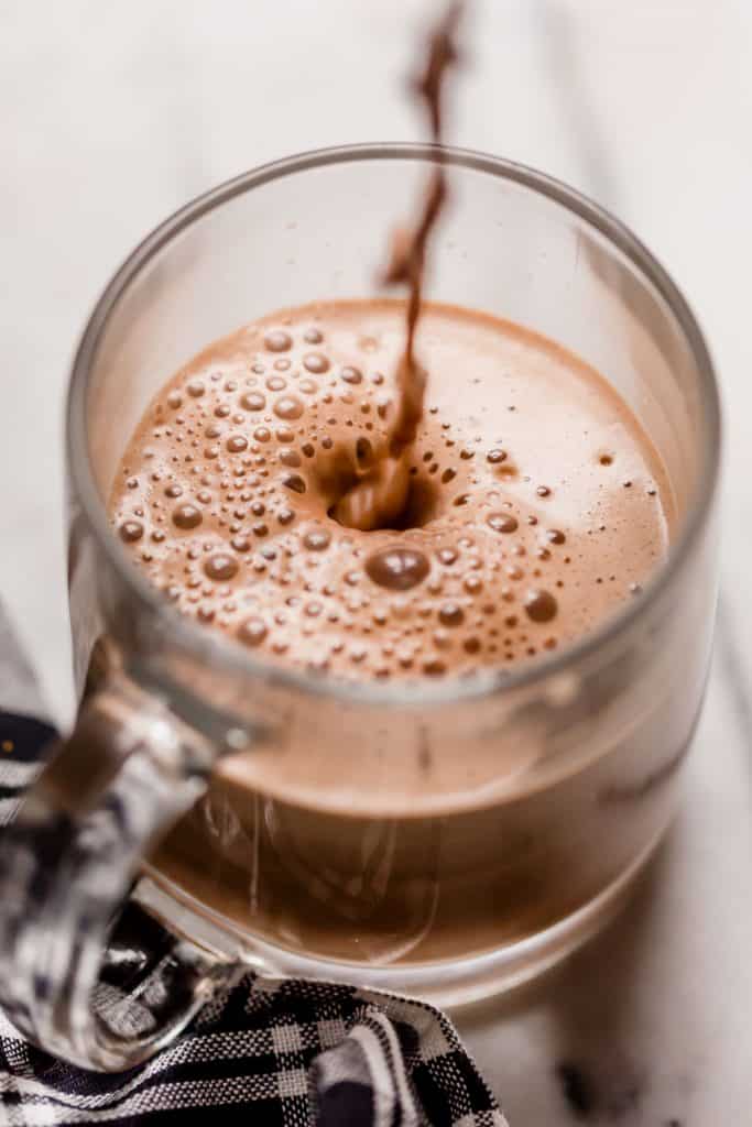 a serving of frothy bulletproof peppermint mocha poured in a clear glass mug on top of a marble countertop next to a black and white checkered linen napkin