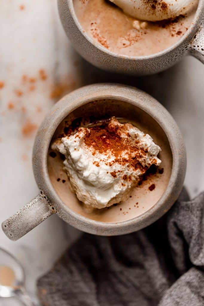 overhead shot of keto cinnamon latte with dollop of whipped cream and sprinkle of cinnamon sitting atop marble countertop with dark linen napkin and metal spoon next to cup