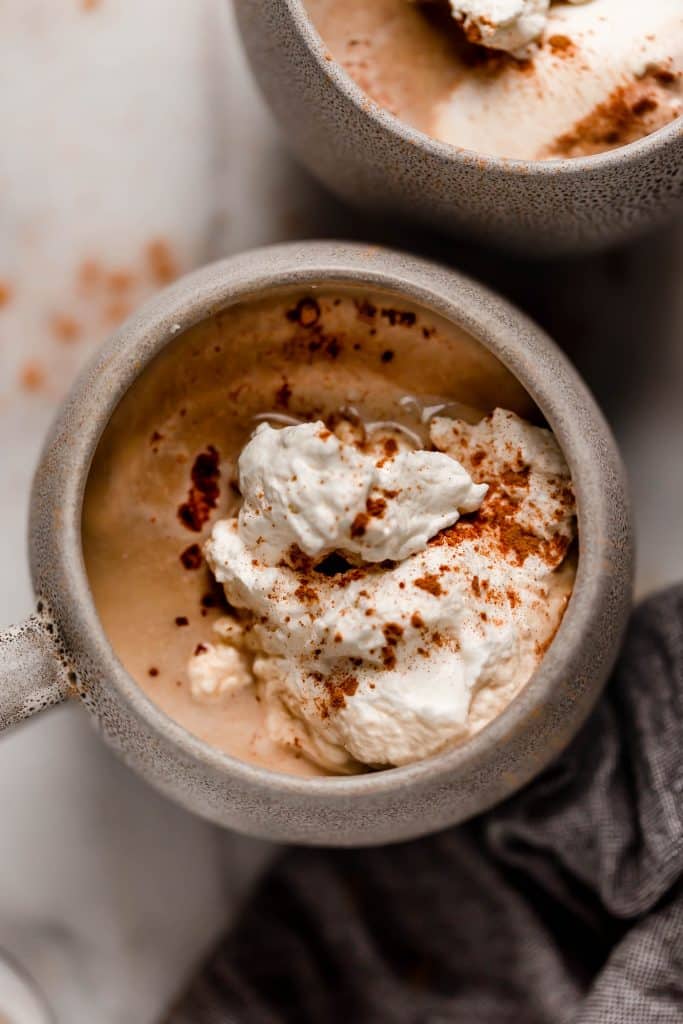 overhead shot of a mug of cinnamon dolce latte with whipped cream and cinnamon on top next to dark cloth linen