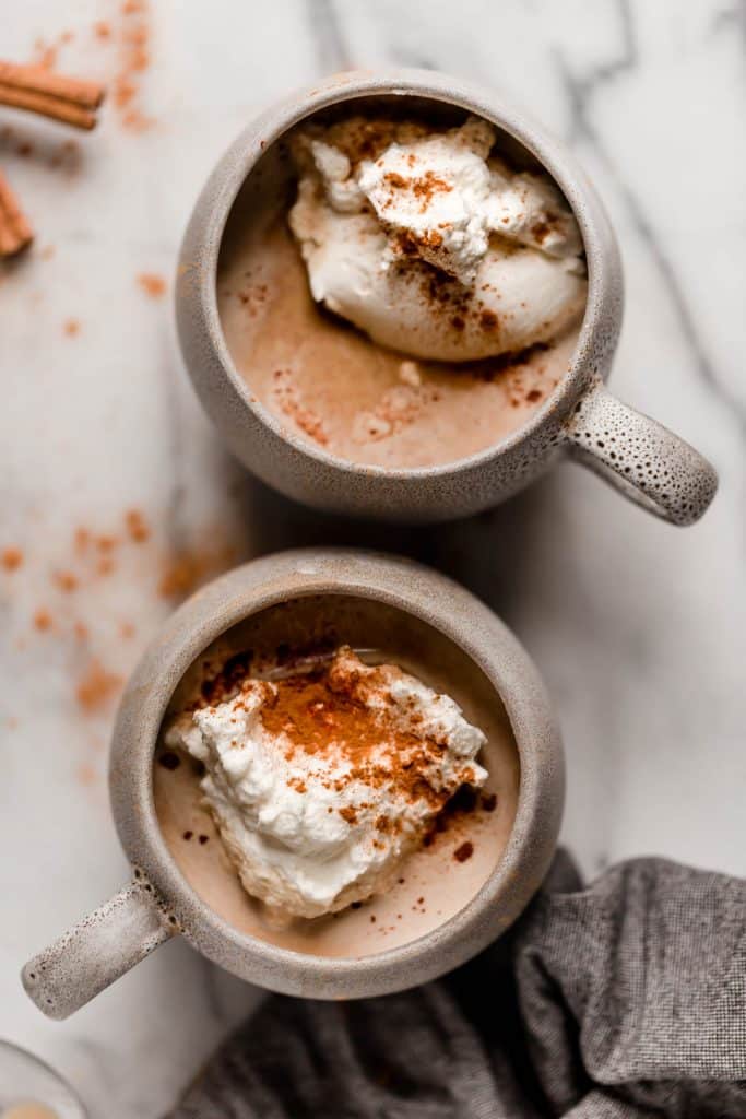 two mugs of latte with whipped cream on top of a marble counter with cinnamon sticks on the side
