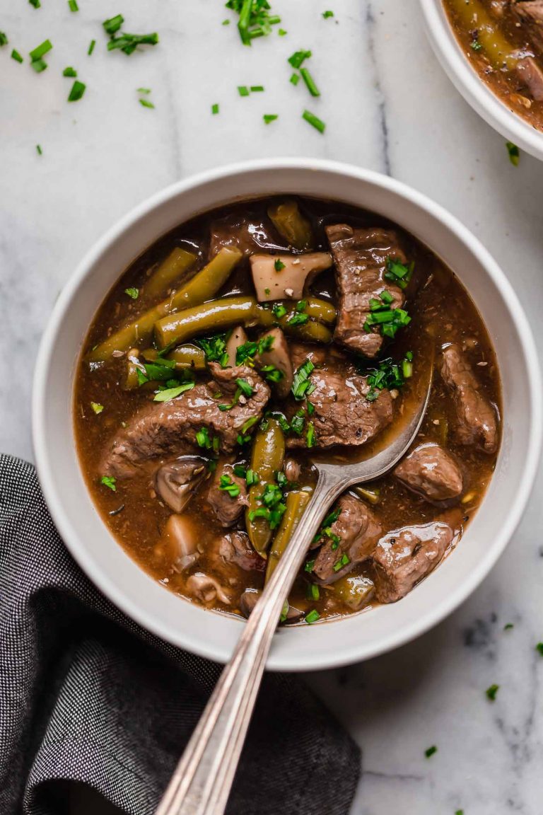 overhead shot of instant pot beef stew in a ceramic bowl with a spoon next to a dark linen napkin on top of a marble countertop