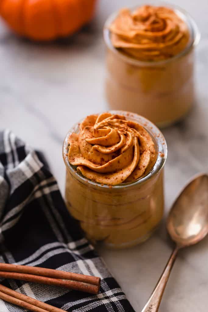 side angle shot of pumpkin mousse sprinkled with cinnamon in glass jars with spoon and cloth napkin next to jar