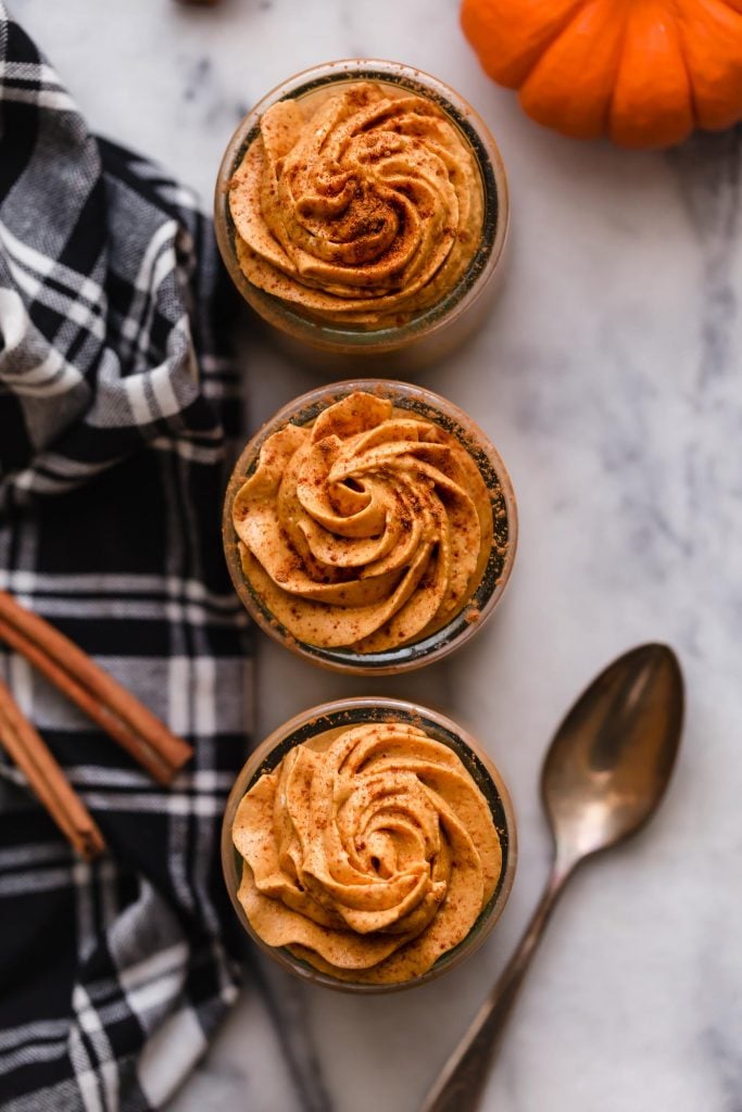 overhead shot of 3 glass jars filled with prepared low-carb pumpkin mousse with cinnamon sprinkled on top next to a spoon, cloth napkin, and cinnamon sticks atop a marble countertop