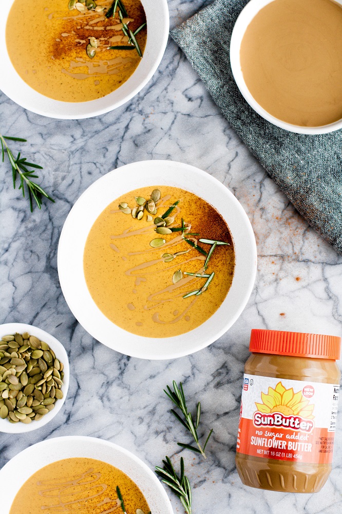 three bowls of pumpkin sunbutter soup garnished with rosemary sprigs with pumpkin seeds for garnishing and a jar of SunButter on the side