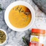 a bowl of pumpkin sunbutter soup beside a small cup of shelled pumpkin seeds and a jar of SunButter
