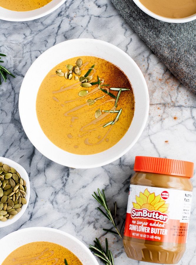 a bowl of pumpkin sunbutter soup beside a small cup of shelled pumpkin seeds and a jar of SunButter