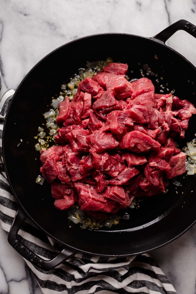 raw chopped beef pieces added onto a large cast iron sauce pan with sauteed onion and garlic