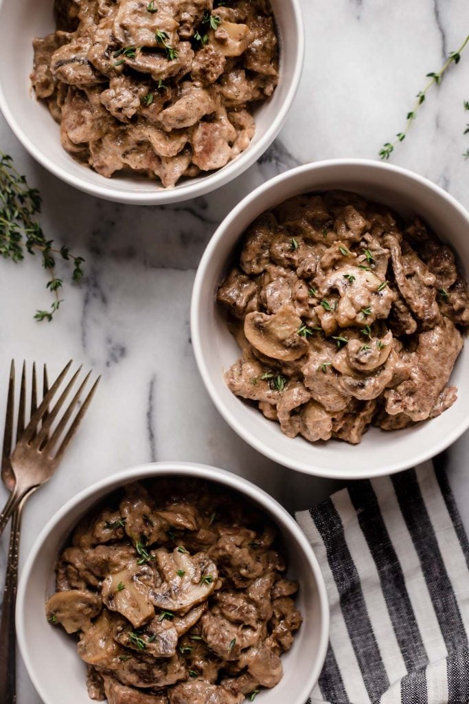 three bowls of keto beef stroganoff atop a marble kitchen counter beside forks