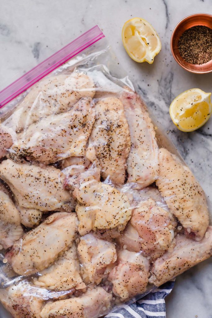 raw marinated lemon pepper chicken wings in a ziplock bag beside two lemon wedges and some seasoning