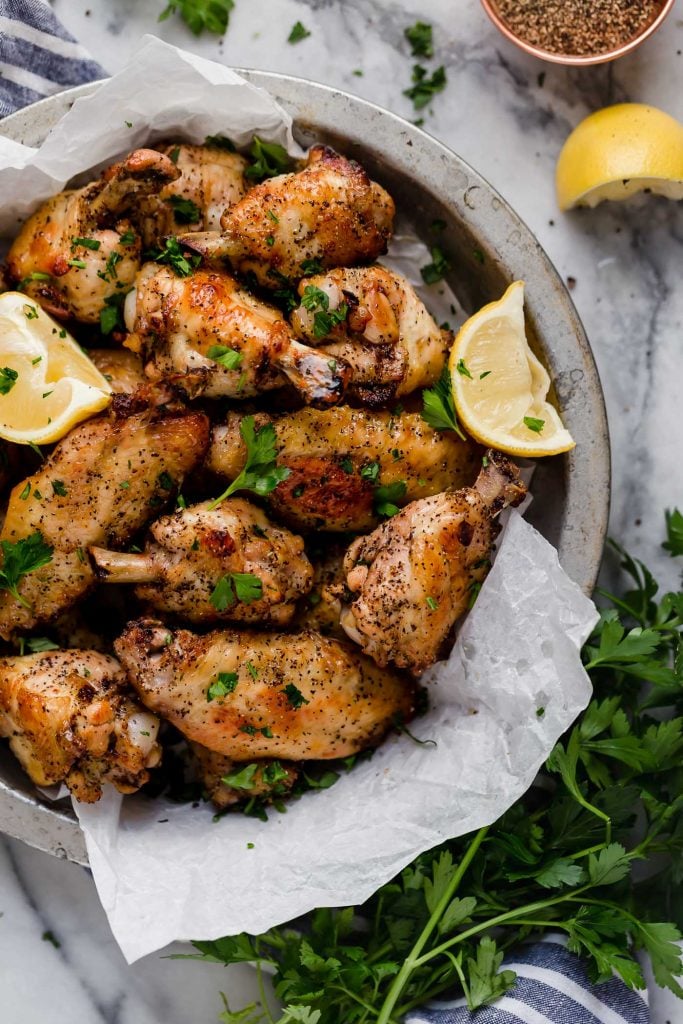 overhead shot of lemon pepper chicken wings in a bowl lined with parchment paper with lemon wedges and parsley