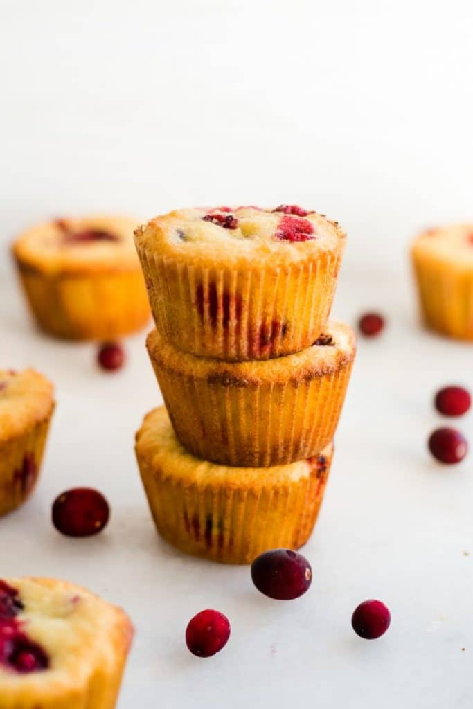 a stack of 3 keto cranberry orange muffins surrounded by fresh cranberries in front of a white background atop a white countertop