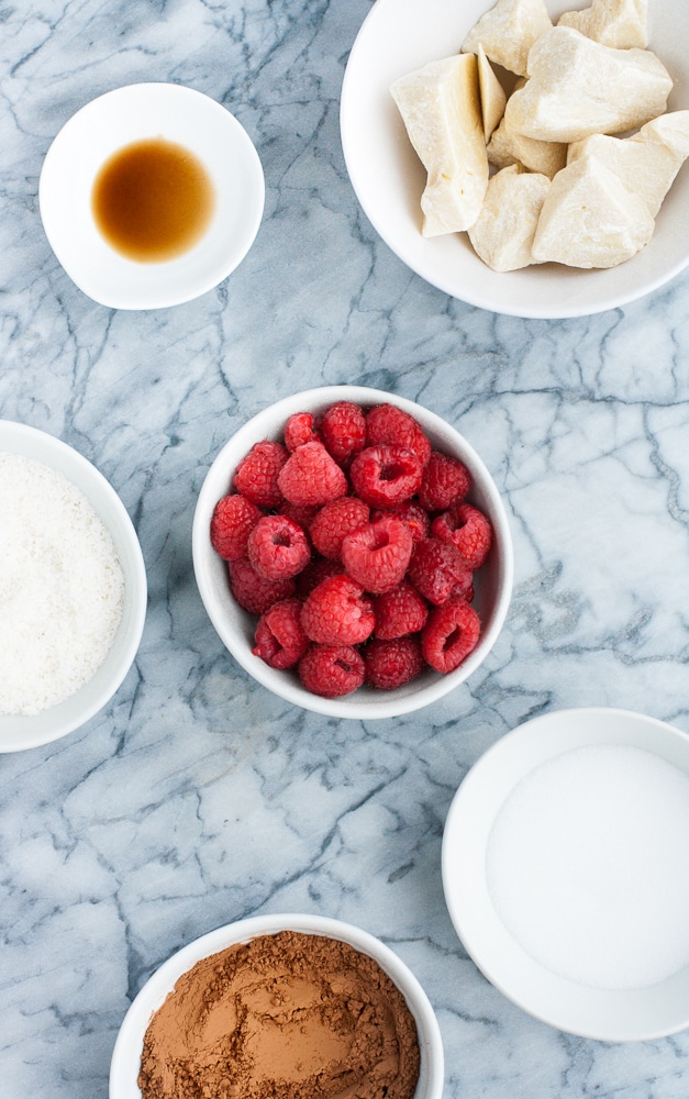 fresh raspberries, cocoa butter, unsweetened cocoa powder, unsweeted shredded coconut, monk fruit sweetener, and pure vanilla extract in small containers atop a marble counter