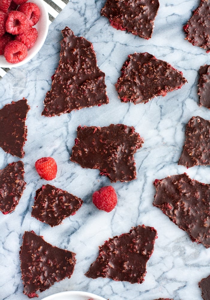 overhead shot of dark chocolate raspberry bark pieces atop a marble counter with fresh raspberries