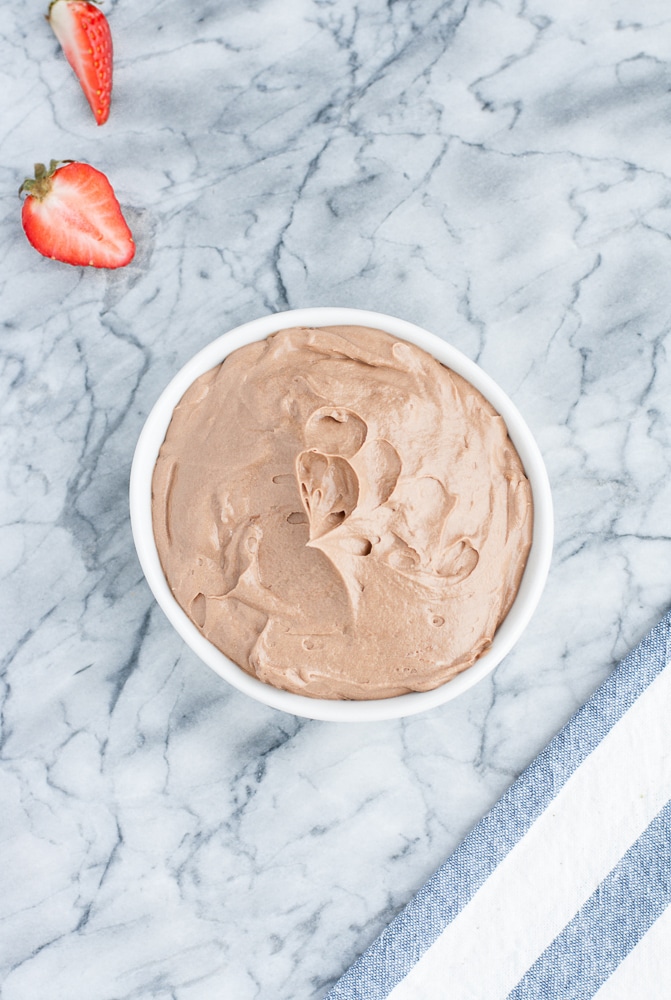 Low Carb Chocolate Strawberry Cupcakes frosting in a ceramic container atop a marble kitchen counter beside a halved strawberry