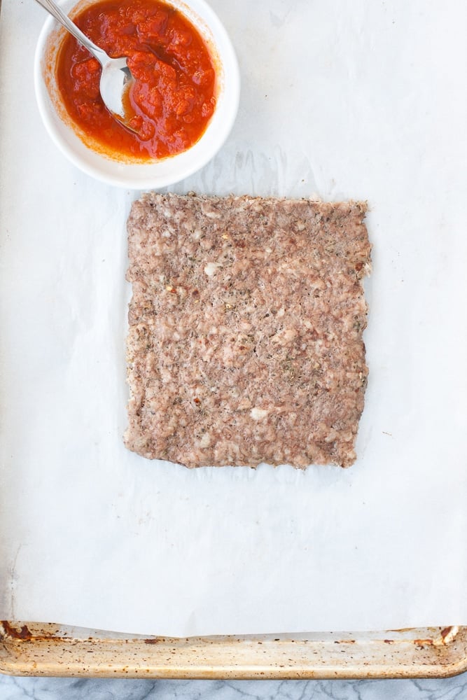 baked ground pork mixture on a sheet of parchment paper beside a bowl of tomato sauce