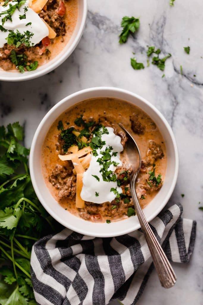overhead shot of a bowl of slow cooker taco soup with a dollop of sour cream, grated cheddar cheese and a sprinkle of fresh parsley with a spoon atop a marble counter with a striped cloth table napkin