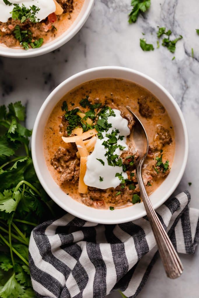 overhead shot of a bowl of slow cooker taco soup with a dollop of sour cream, grated cheddar cheese and a sprinkle of fresh parsley with a spoon atop a marble counter beside a striped cloth tablenapkin