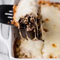 Angled close-up shot of the Low-Carb Philly Cheesesteak Casserole on a baking dish with a serving scooped up using a spatula. The baking dish sits atop a marble countertop.