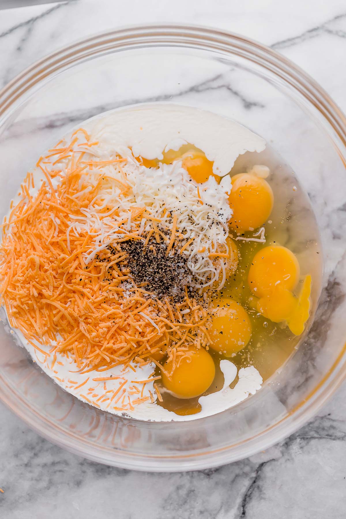 eggs, shredded cheddar, shredded mozzarella, heavy cream, salt, pepper on a glass mixing bowl