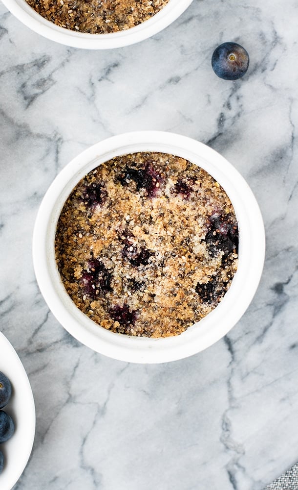 baked blueberry crumble in a white ceramic ramekin beside a fresh blueberry atop a marble counter top