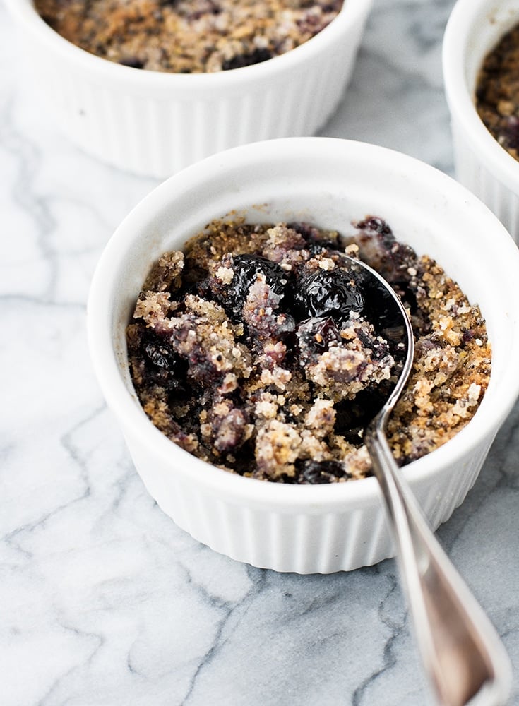 angled shot of keto blueberry crumble in ramekins with a spoon atop a marble kitchen counter