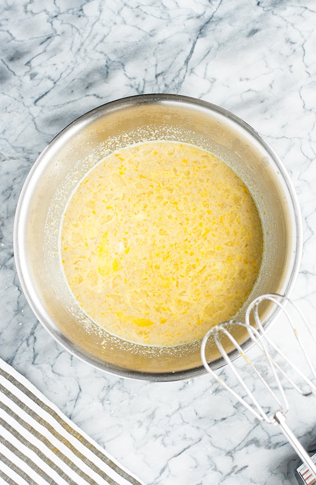 stainless steel mixing bowl with sunflower seed meal, eggs, lemon, juice, and lemon zest mixed together using an electric mixer atop a marble countertop