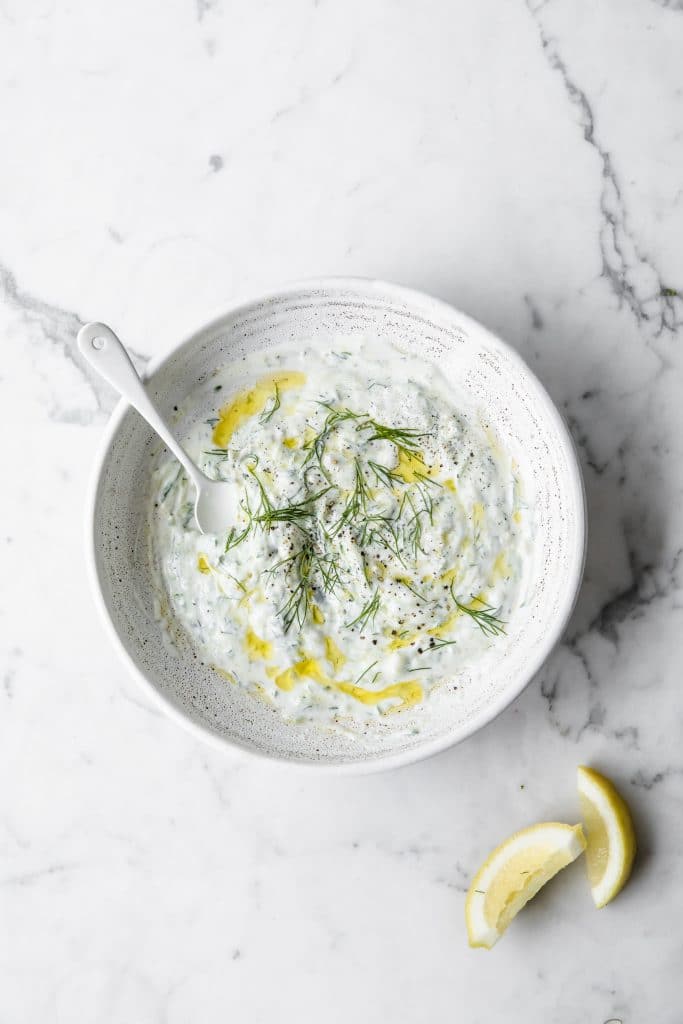 a bowl of keto tzatziki with a teaspon beside lemon wedges atop a marble kitchen counter