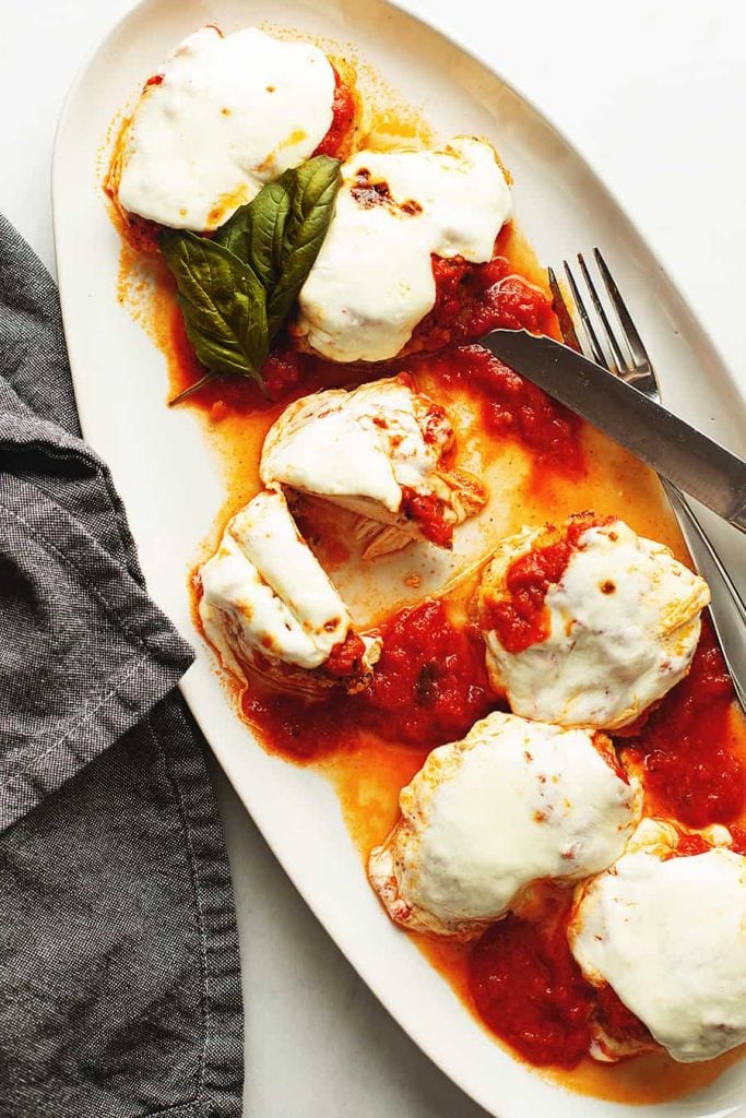 keto chicken parmesan pieces spread on a white serving dish with utensils and basil