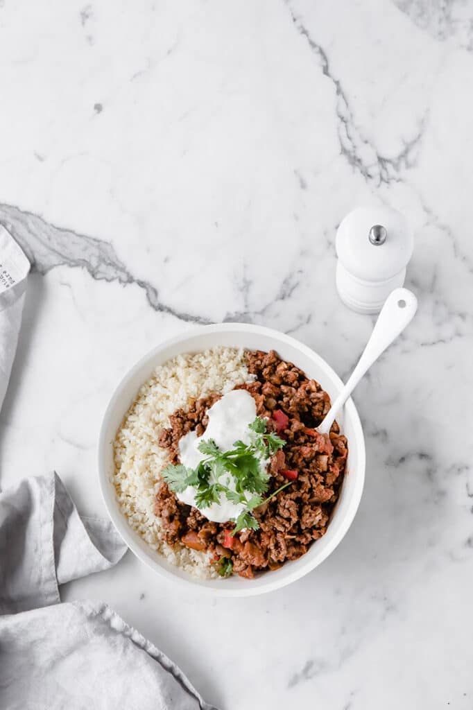 overhead shot of a bowl of keto chili con carne and cauliflower rice atop a marble kitchen counter