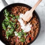 close-up overhead shot of keto chili con carne on a pan topped with sour cream with a wooden spoon atop a marble kitchen counter