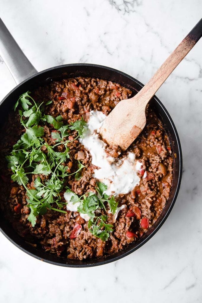 close-up overhead shot of keto chili con carne on a pan topped with sour cream with a wooden spoon atop a marble kitchen counter