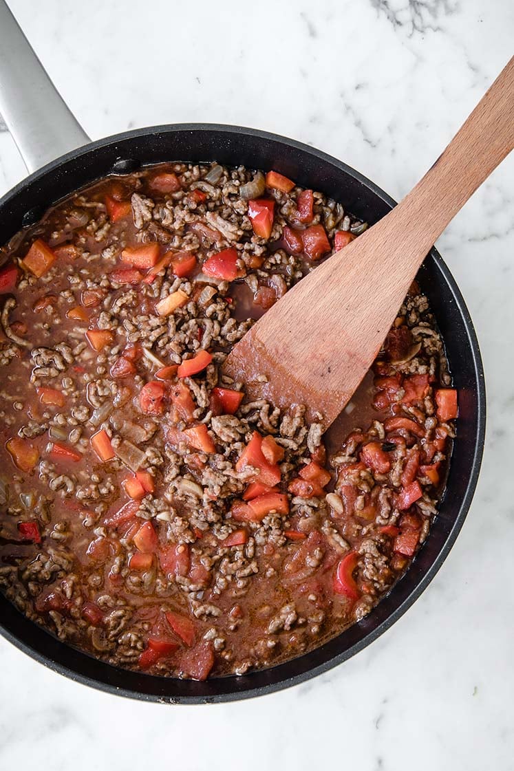 tomato puree poured over ground beef while being stirred in a large pan with a wooden spoon