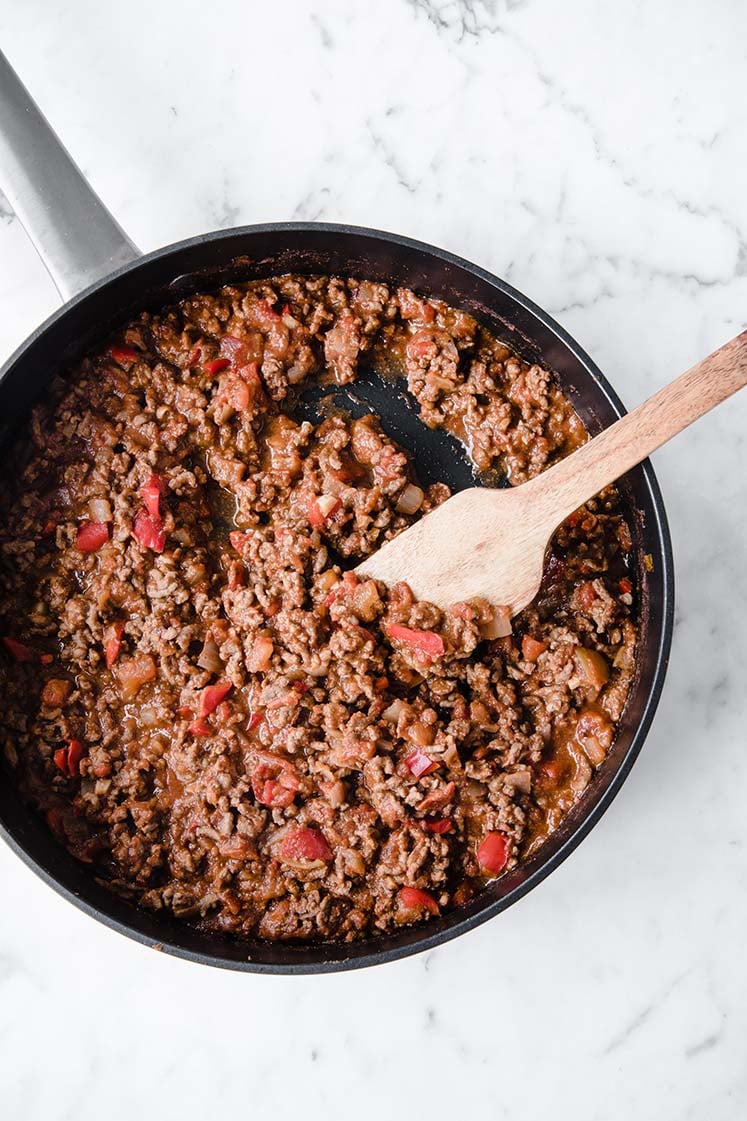 keto hili con carne being simmered until sauce thickens while being stirred with a wooden spoon in a large pan atop a marble kitchen counter