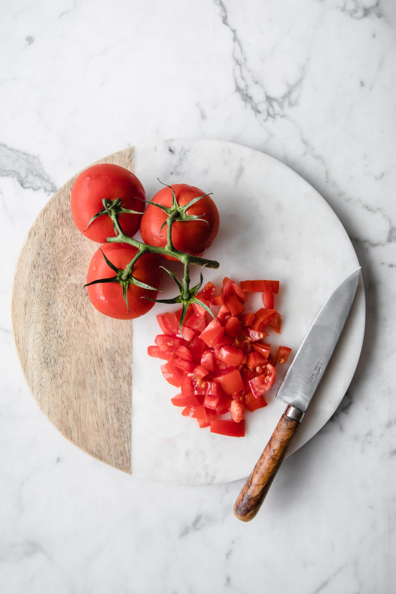 3 whole tomatoes and some diced tomatoes on a chopping board with a knife at the side atop a marble kitchen counter