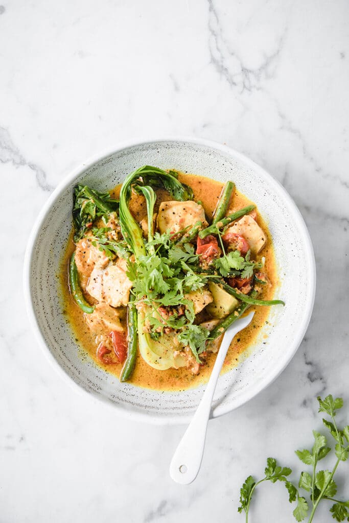 a bowl of keto salmon curry with herbs at the side atop a marble kitchen table