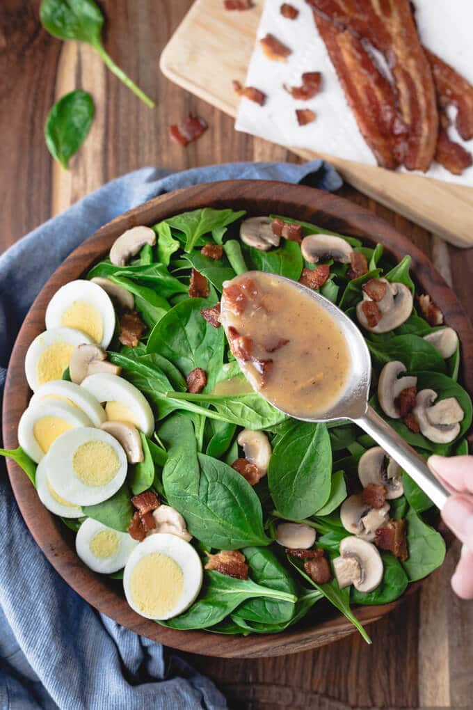 close up shot of spinach salad being drizzled with war bacon dressing atop a wooden table