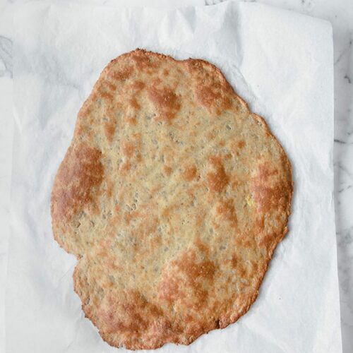 Overhead shot of baked keto pizza crust without almond flour on parchment paper atop a marble kitchen counter.