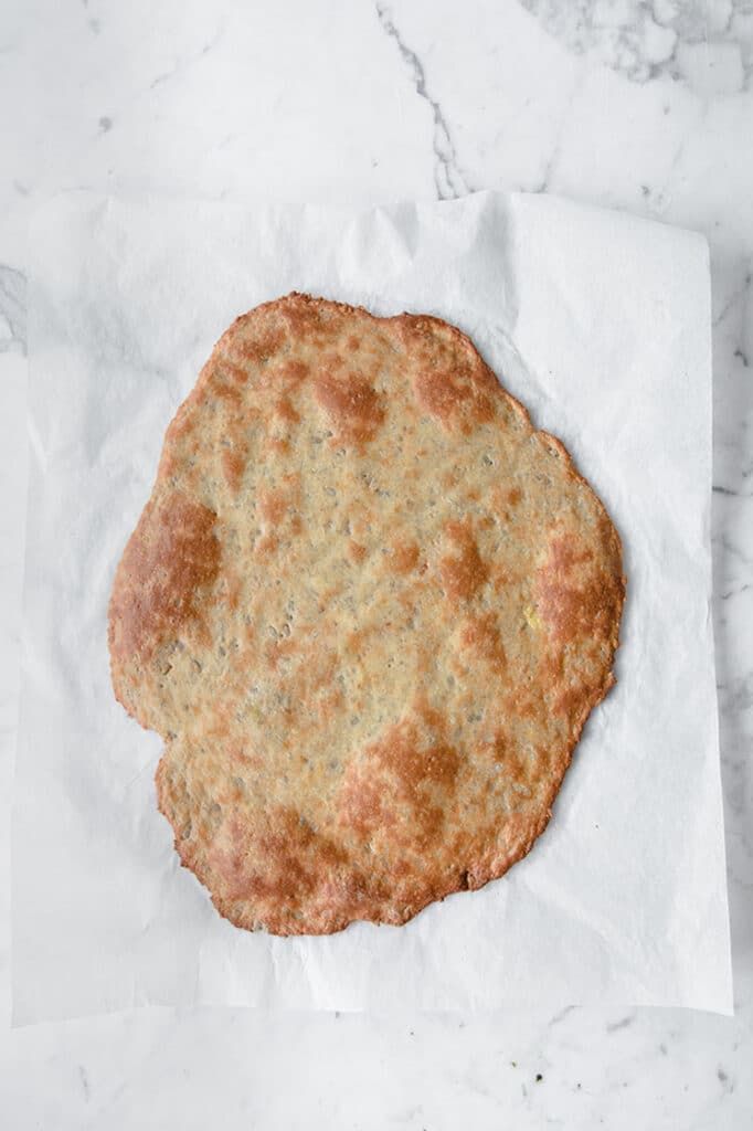 Overhead shot of baked keto pizza crust without almond flour on parchment paper atop a marble kitchen counter.