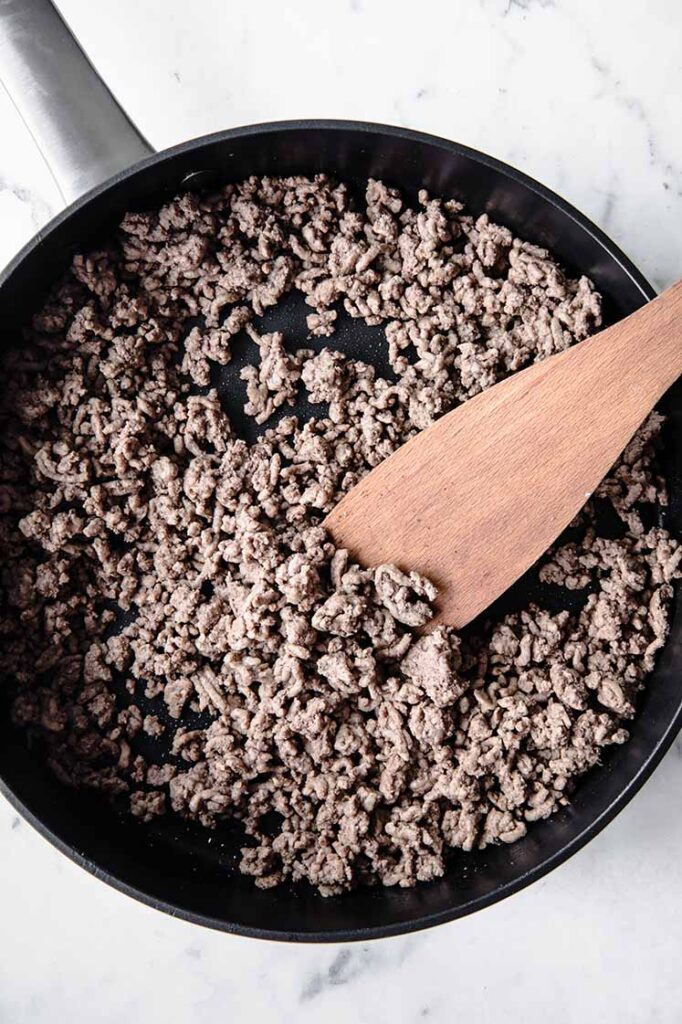 overhead shot of ground beef on a pan with a wooden spatula at the side atop a marble kitchen table counter