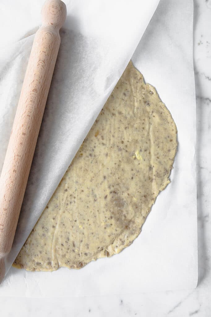 pizza dough flattened using a rolling pin on parchment paper atop a marble kitchen counter