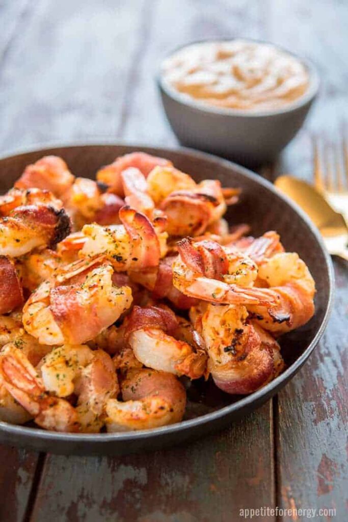 a plate of bacon wrapped shrimp with remoulade sauce atop a wooden table