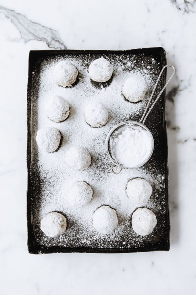 nut-free keto snowball cookies being dusted by powdered sweetener on a baking tray atop a marble kitchen counter