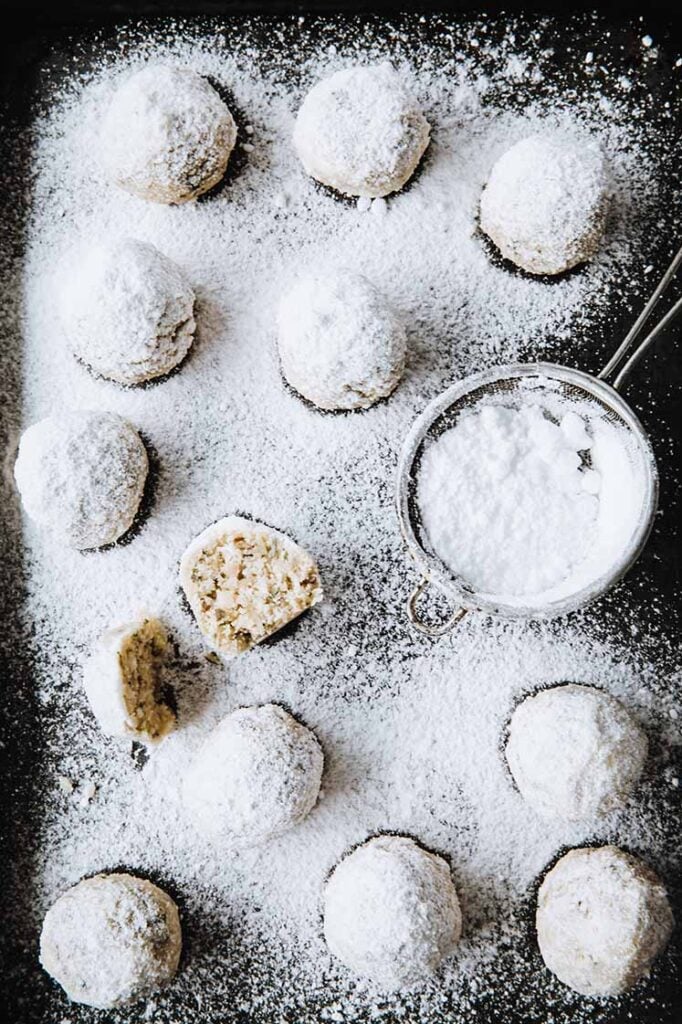 nut-free keto snowball cookie broken in half being dusted by powdered sweetener on a baking tray atop a marble kitchen counter