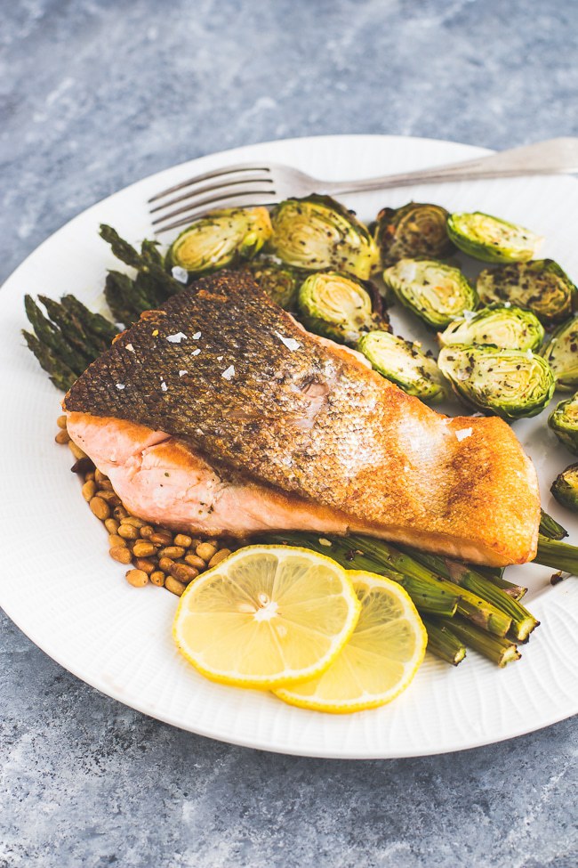 a plate of salmon with asparagus and pine nuts garnished with lemons atop a marble kitchen counter