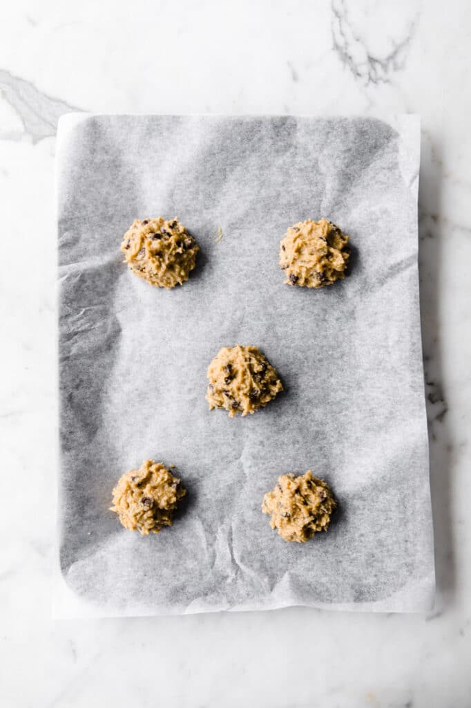 5 nut-free chocolate chip cookie dough rolled into balls on a baking sheet lined with parchment paper atop a marble kitchen table