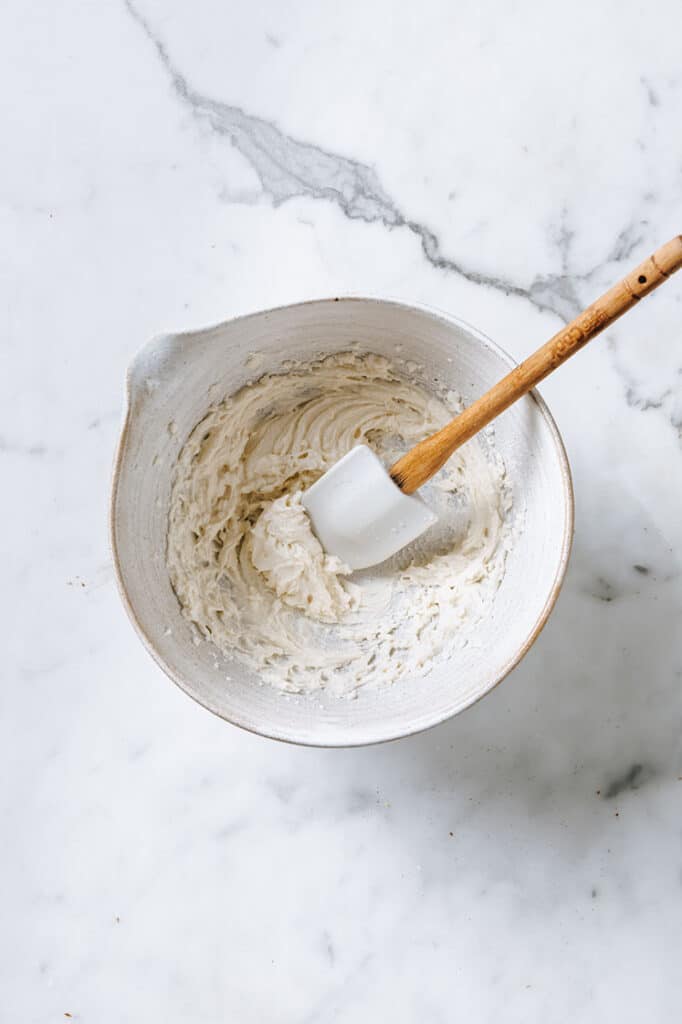 cinnamon roll chaffle icing being stirred with a spatula in a mixing bowl atop a marble kitchen counter