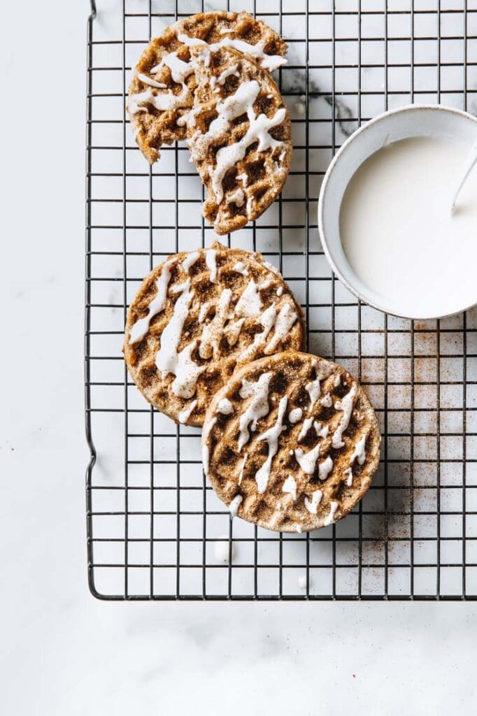 three cinnamon roll chaffles with one sliced in half on a wire rack with a bowl of icing atop a marble kitchen counter